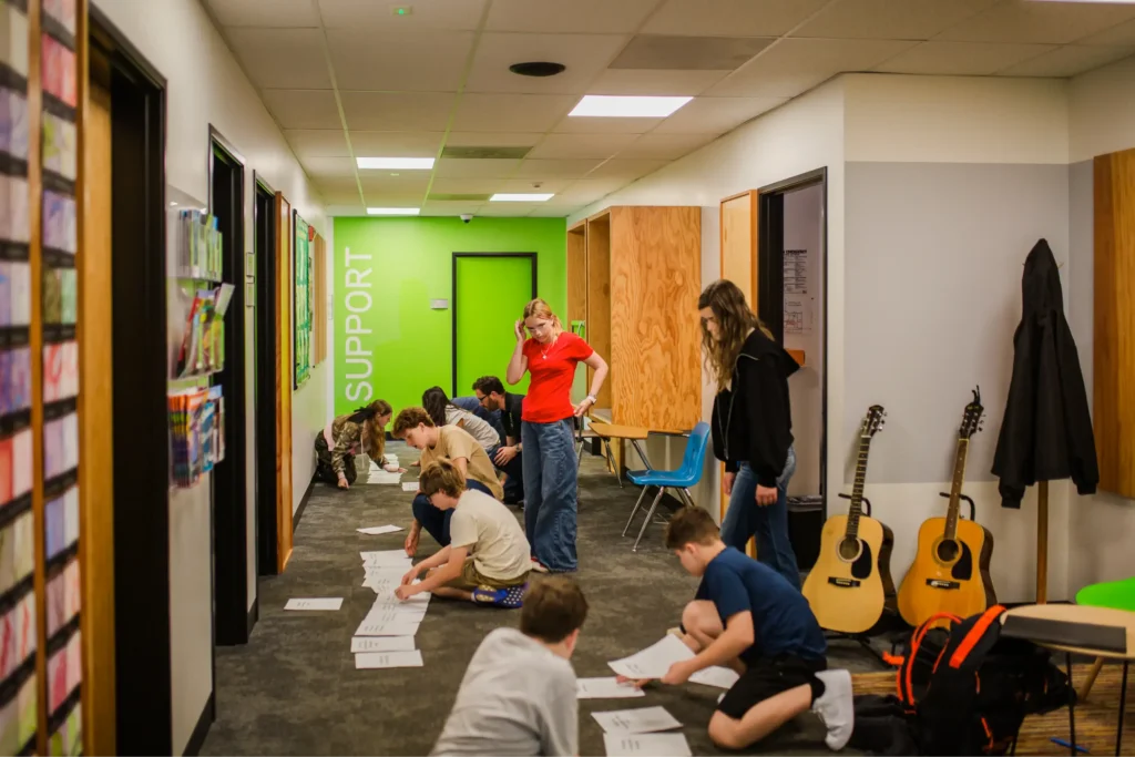 Student working on floor, some seated, some standing, acoustic guitars in the background